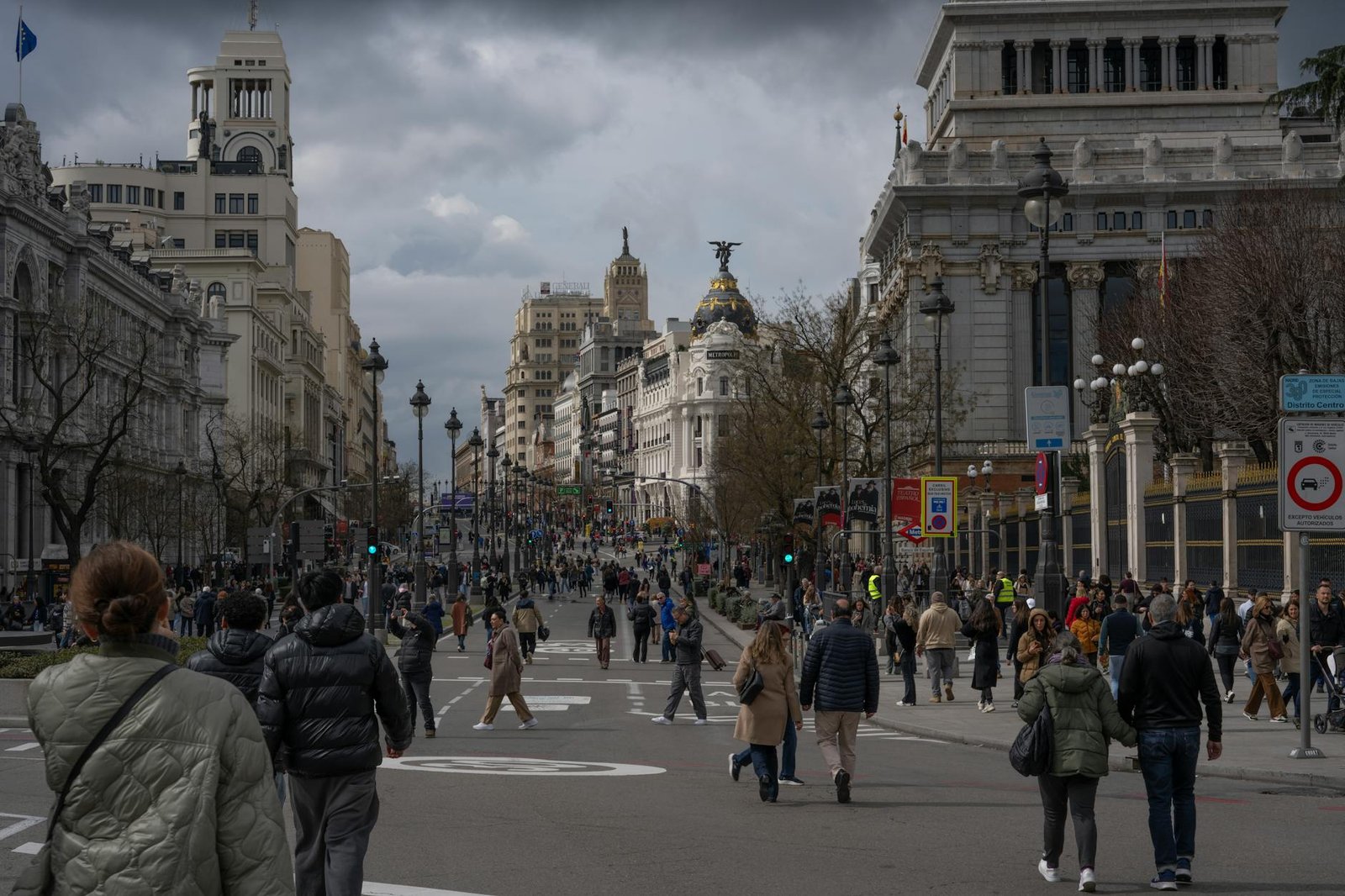 Busy street scene in Madrid's city center with iconic architecture and bustling pedestrians under a cloudy sky.