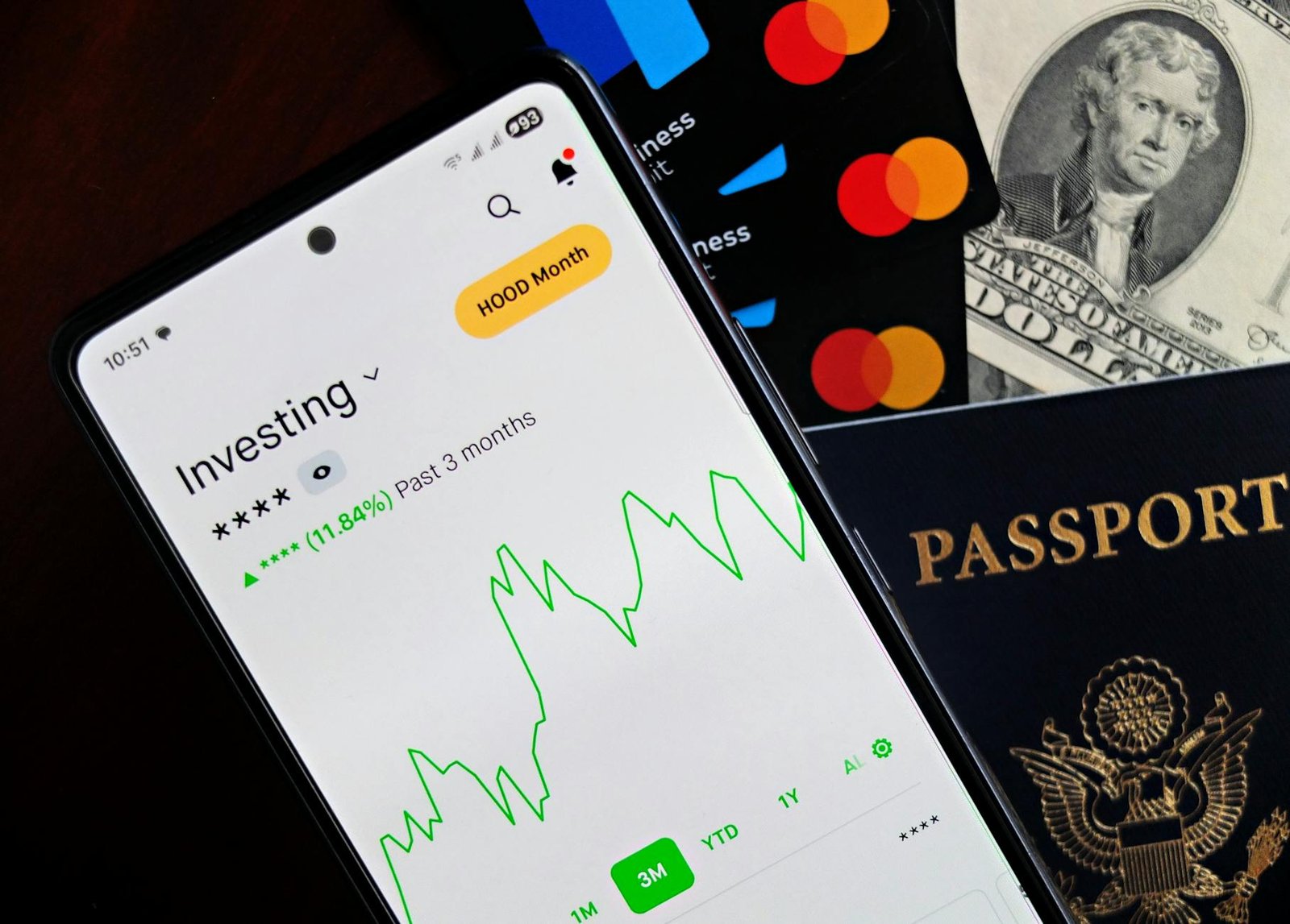 Smartphone displaying investing app, with credit cards, cash, and passport nearby, symbolizing finance